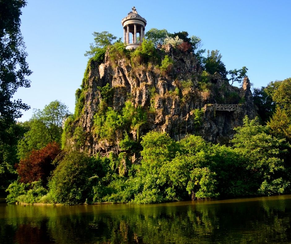 A small temple sits on top of a craggy hill at the edge of a lake in arc des Buttes-Chaumont